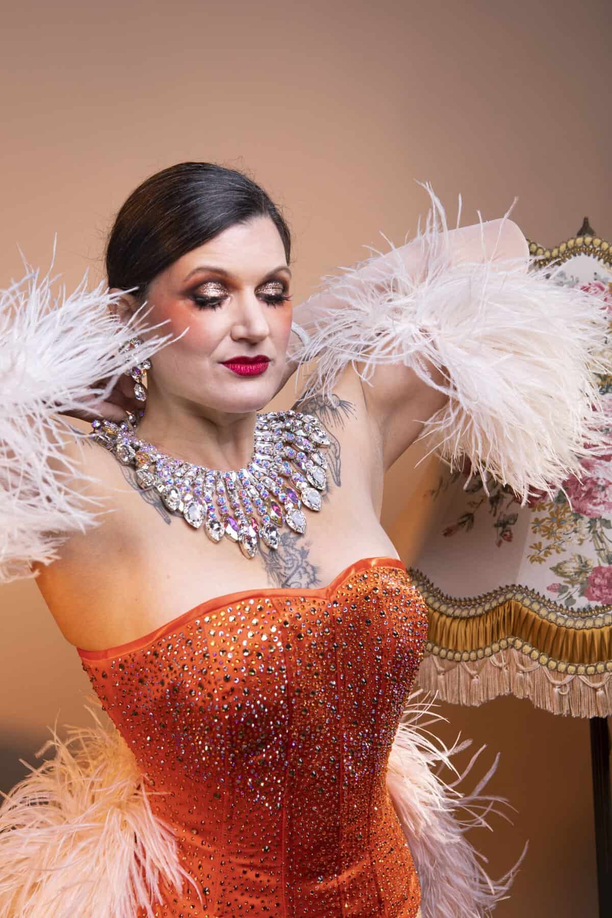Une femme vêtue d'un corset orange étincelant avec des détails en plumes pose avec ses mains près de son visage. Elle porte un maquillage spectaculaire, un collier de bijoux et se tient à côté d'une lampe à motifs floraux.