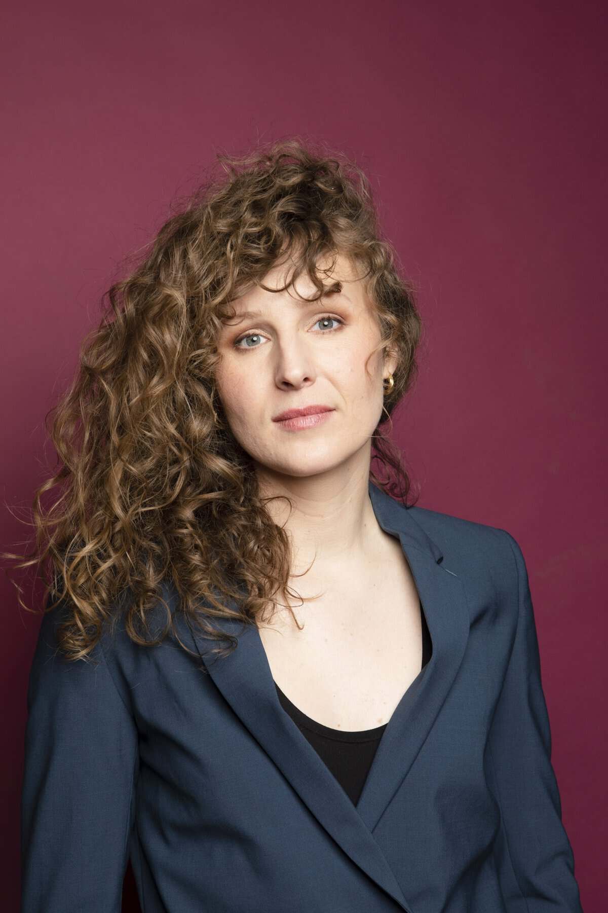 Une personne aux longs cheveux bruns bouclés porte un blazer bleu sur un haut noir et pose sur un fond marron uni. Elle a une expression neutre et regarde légèrement vers l'appareil photo.